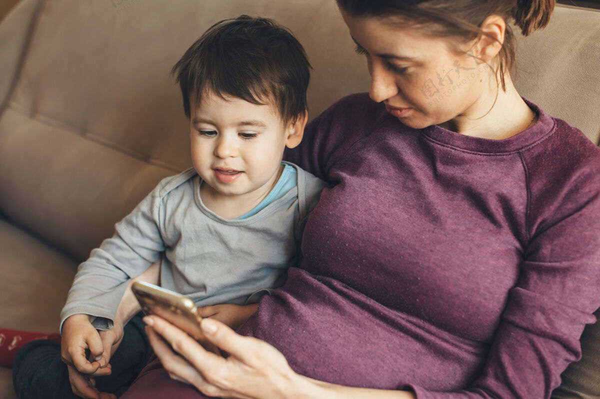 父母怀孕的白人妇女和她的小儿子坐在沙发上看电话等待儿子应用程序