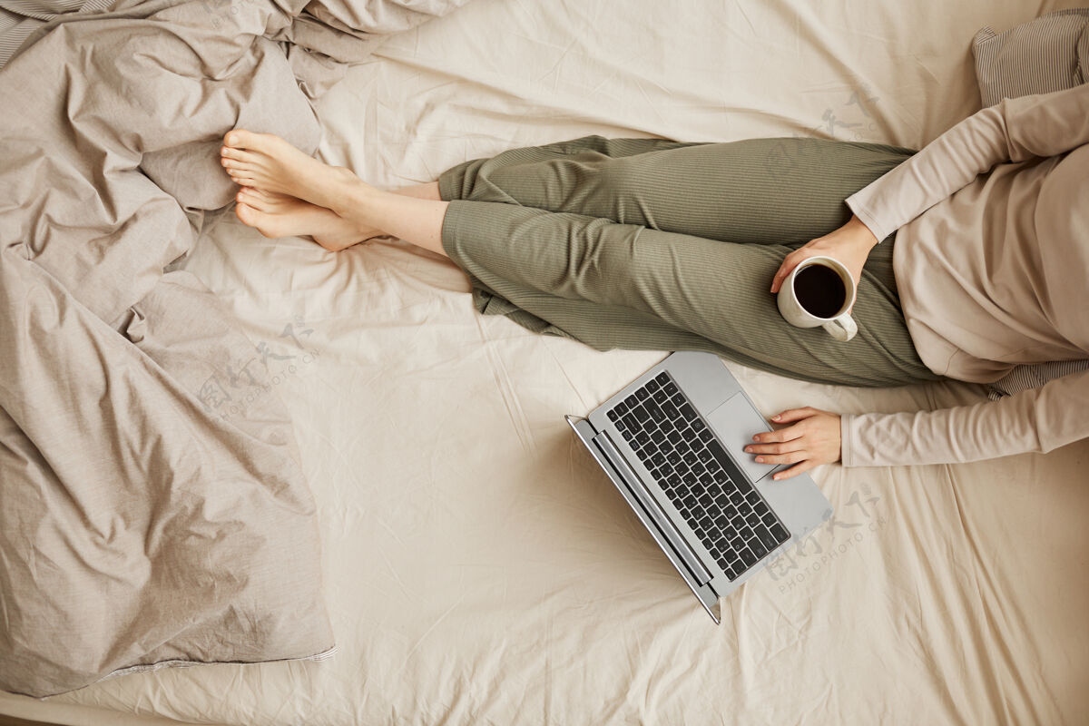 睡衣一个女人坐在床上喝着咖啡 早上用笔记本电脑的特写镜头家庭室内躺下床