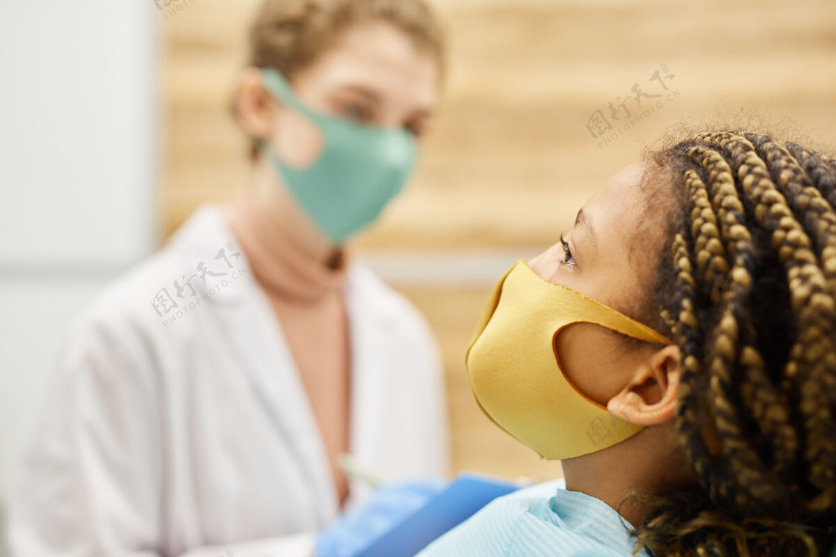 成人戴着防护面具的小女孩躺在医院的侧视图 医生在墙上制服牙科设备职业
