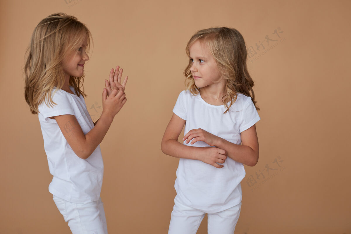 快乐正面是一对可爱的小双胞胎姐妹的肖像 她们卷曲着金色的头发 互相交谈 穿着白色的衣服横着看朋友女孩时尚