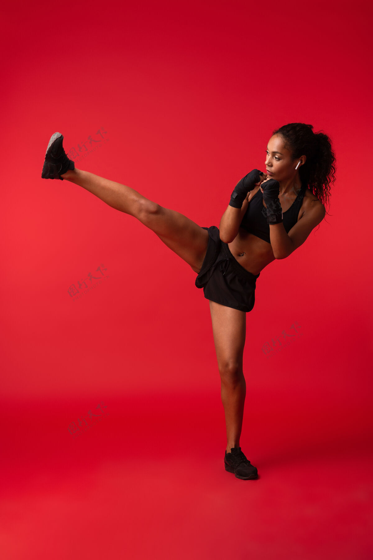 健美操穿着黑色运动服拳击和踢腿的运动型非裔美国妇女的肖像 隔离在红墙外拳击伸展墙壁