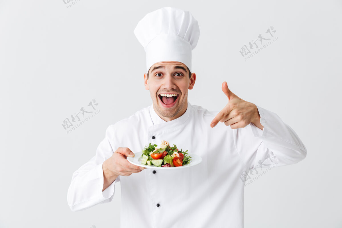 手兴奋的男厨师穿着制服 把新鲜的蔬菜沙拉放在隔离在白色墙上的盘子上专业吸引力烹饪