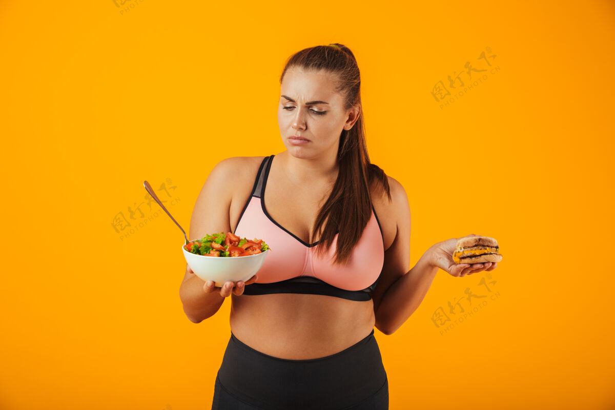 休闲一个心烦意乱的超重健身妇女穿着运动服 孤立地站在黄色的墙上 手里拿着盛着沙拉和汉堡的碗健身女孩护理