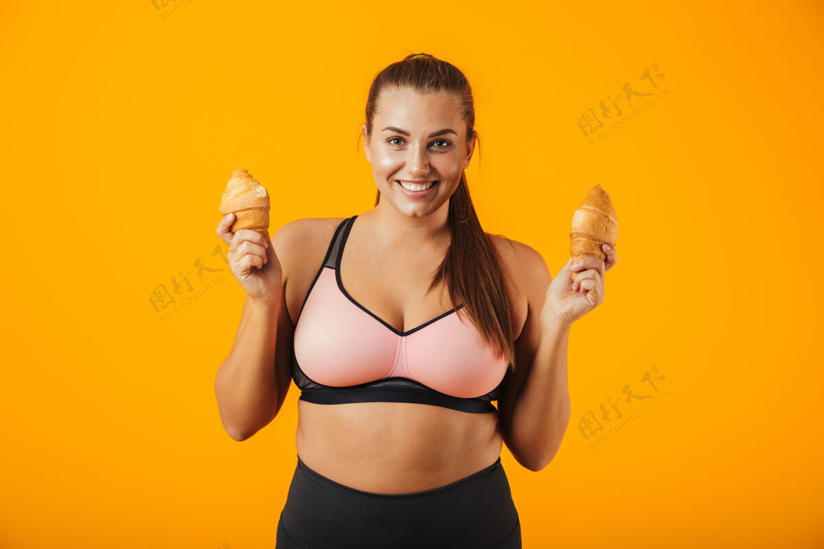 运动装一个穿着运动服的快乐大女人 双手拿着两个牛角包 隔离在黄色背景下女性羊角面包运动型