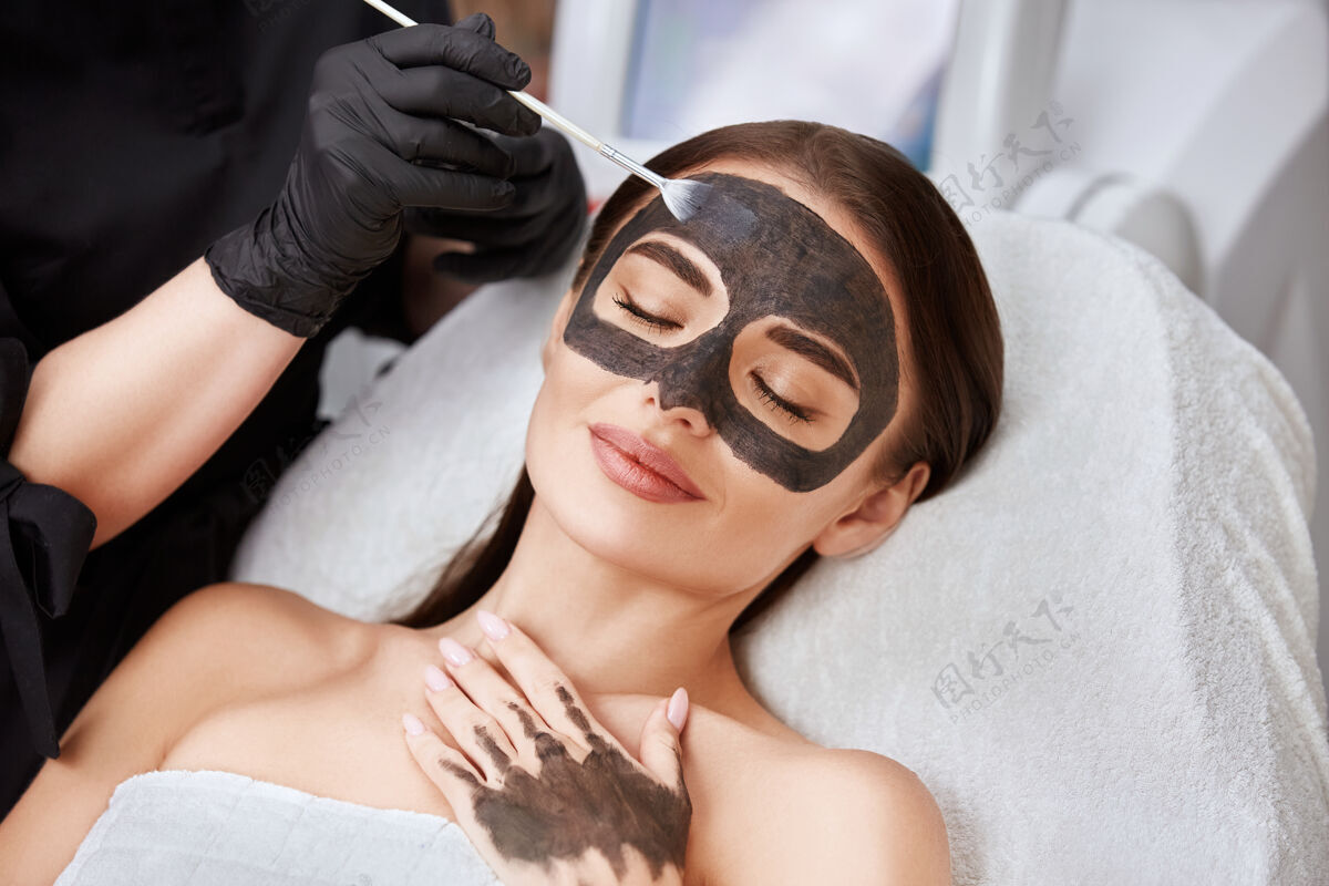 面部用碳素面膜和美容师手臂贴出的女人脸特写 可爱的女人正在接受美容师的面部护理化妆品粘土面部