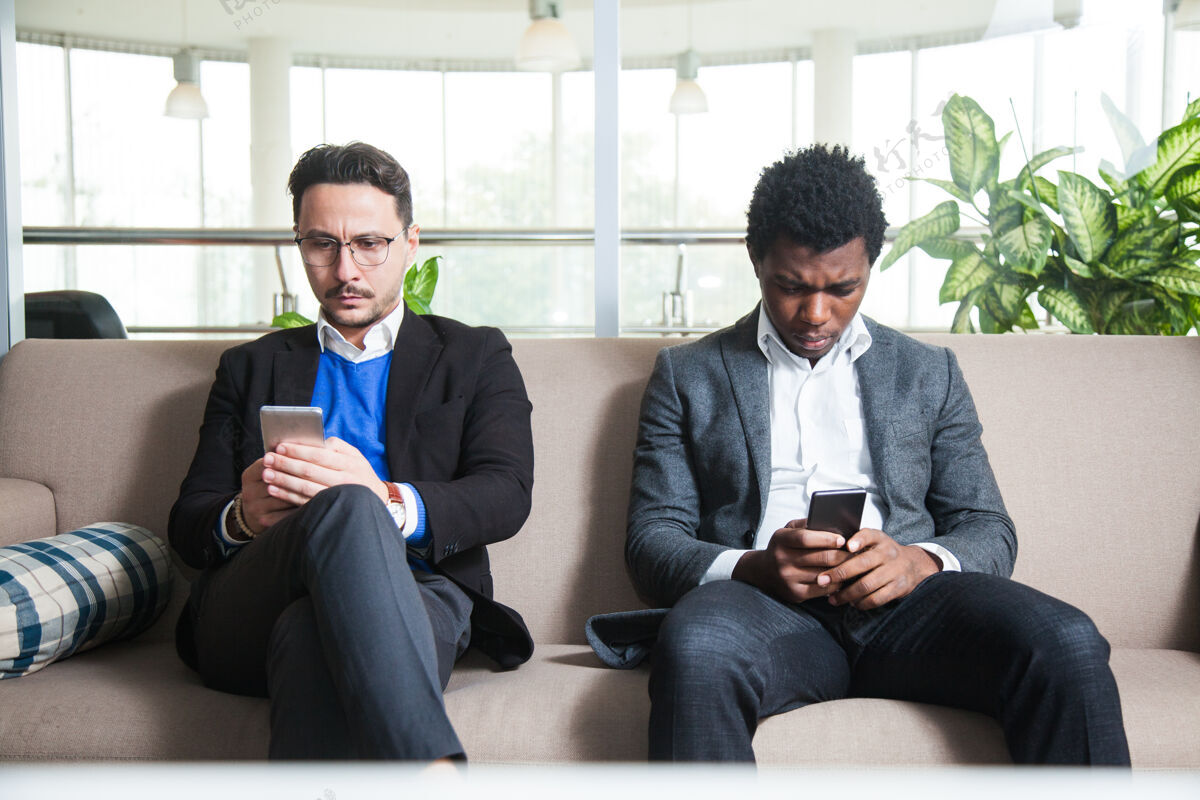 消息两个多元文化的男人坐在沙发上拿着手机短信通信网络