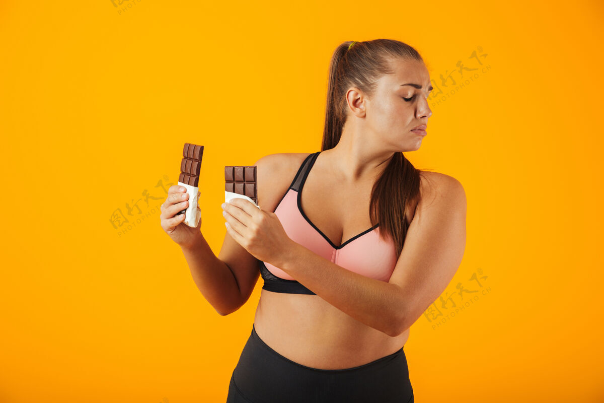 女人一个穿着运动服的心烦意乱的超重年轻女子的肖像 孤立地站在黄色的墙上 手里拿着巧克力棒饮食站身体