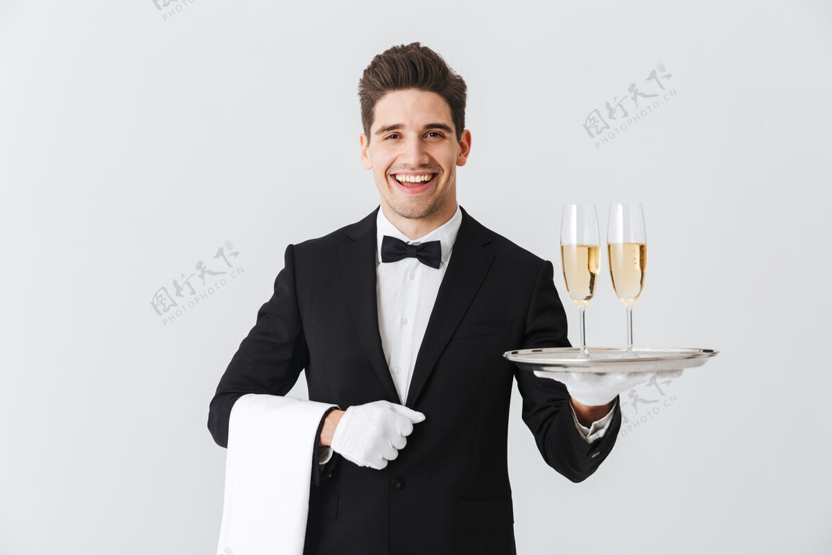 前面一个穿着燕尾服的微笑的年轻侍者的肖像 白色墙壁上放着两杯香槟男性工作饮料