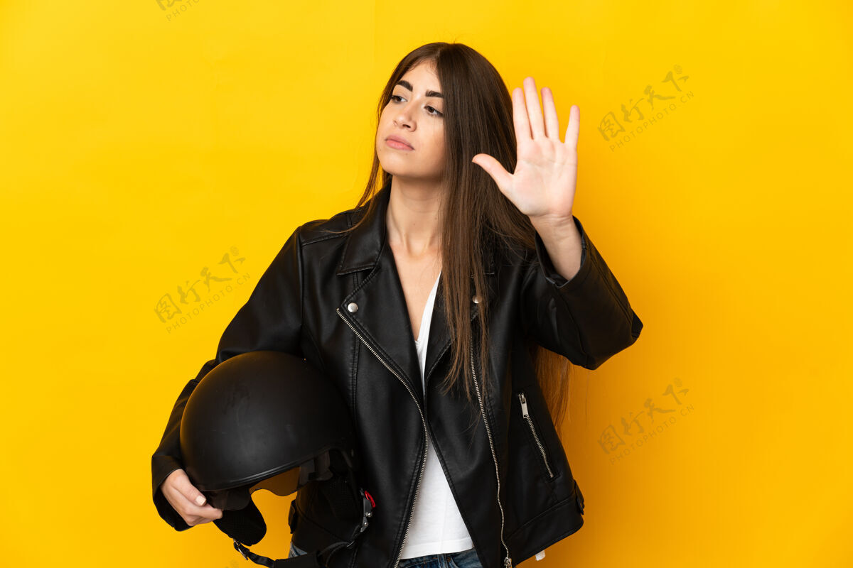人年轻的高加索女子手持摩托车头盔孤立在黄色背景下做停车手势和失望手势控制驾驶