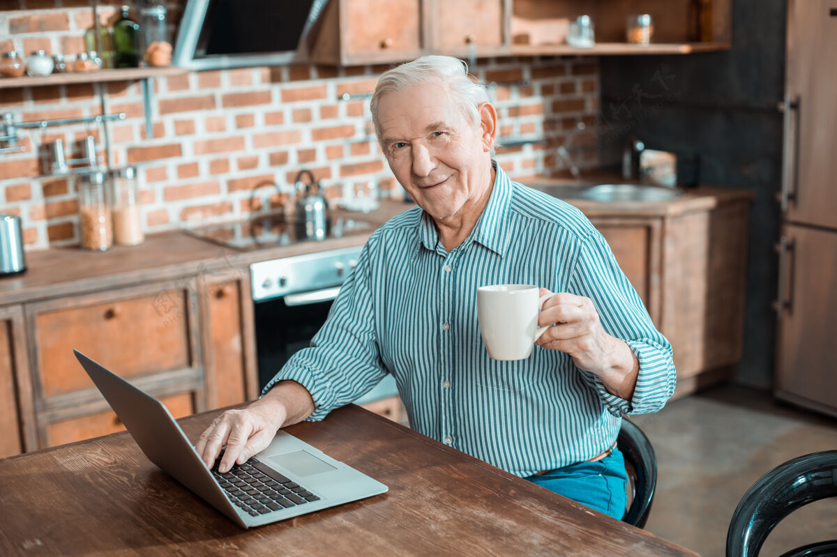 退休开朗积极的老人端着一杯茶 一边看着你一边用笔记本电脑工作笔记本电脑休息休闲