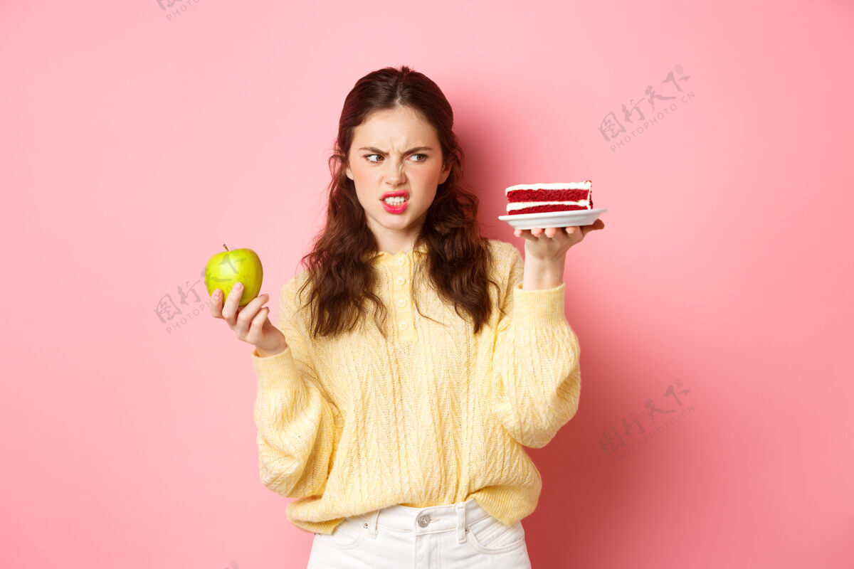 水平节食的健康女孩厌恶地看着一块蛋糕 拿着绿色的敷 做着不满意的鬼脸 站在粉红色的墙上积极乐观蛋糕