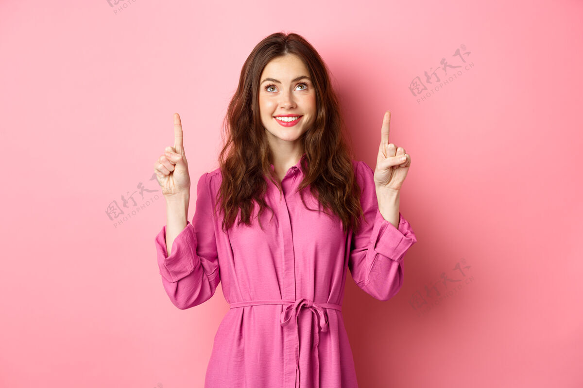 女人美丽快乐的女士穿着礼服 指指点点 面带微笑 面带喜悦 盯着宣传文字 读着你的标志 站在粉红色的墙上时尚欢呼兴奋