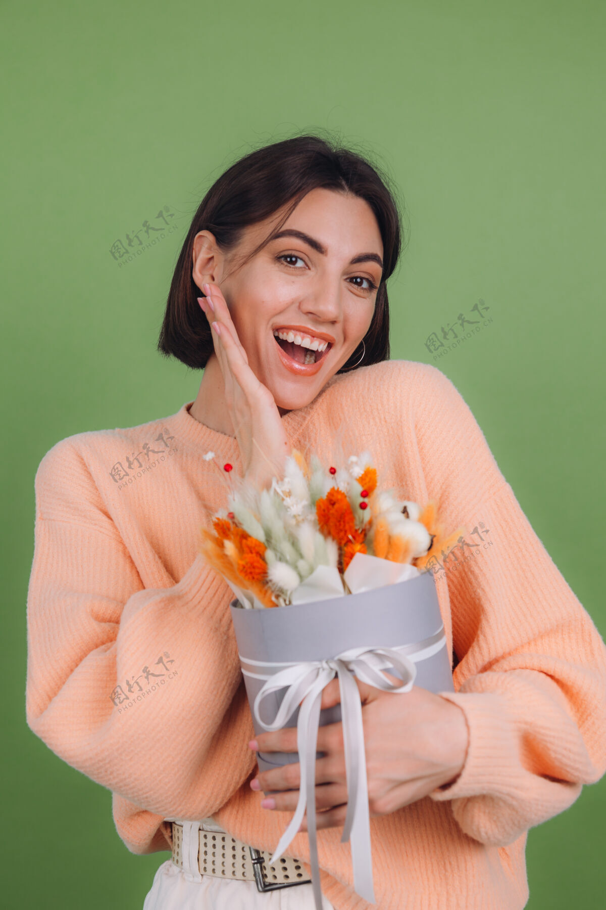 礼盒年轻女子穿着休闲桃色毛衣隔离在绿橄榄墙上手持橙白色花盒组成的棉花 吉普赛拉小麦和拉古鲁斯作为礼物开心惊喜吉普赛拉女性棉花花