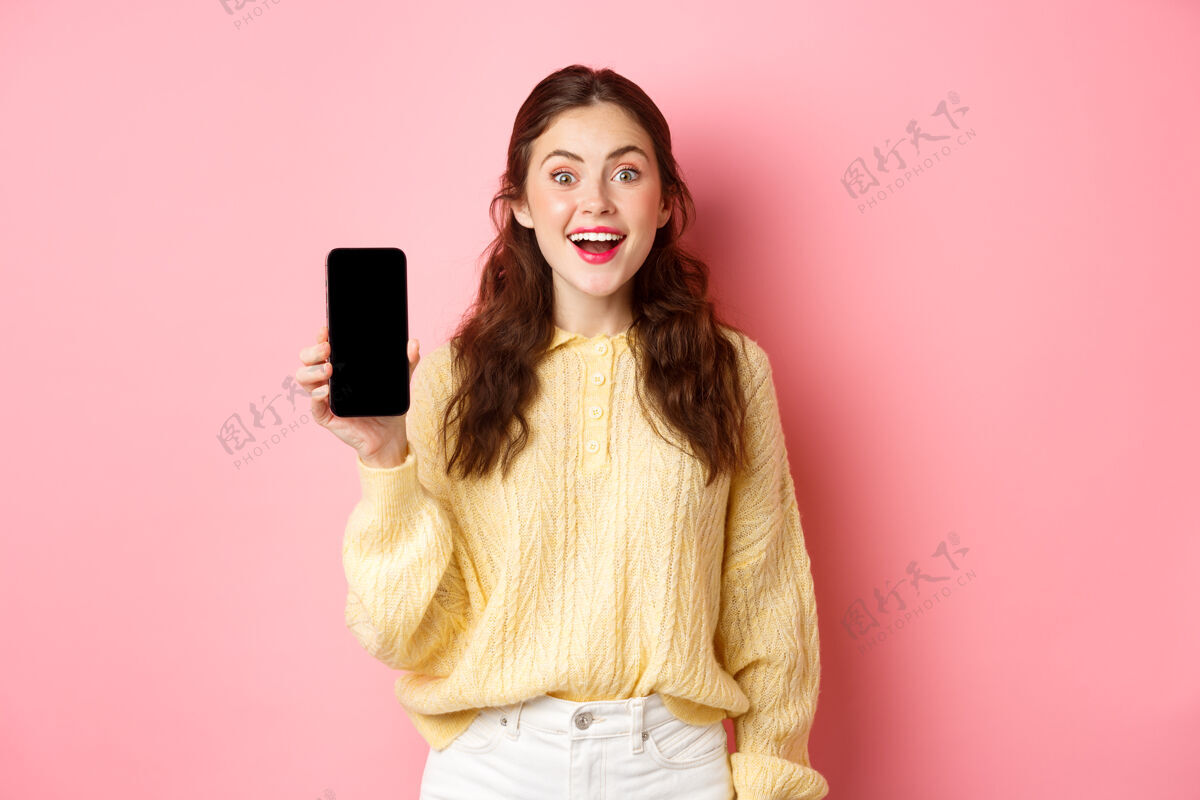 情绪科技和网上购物迷人的女学生看起来很兴奋 显示空的智能手机屏幕和微笑 站在粉红色的墙壁在线兴奋魅力