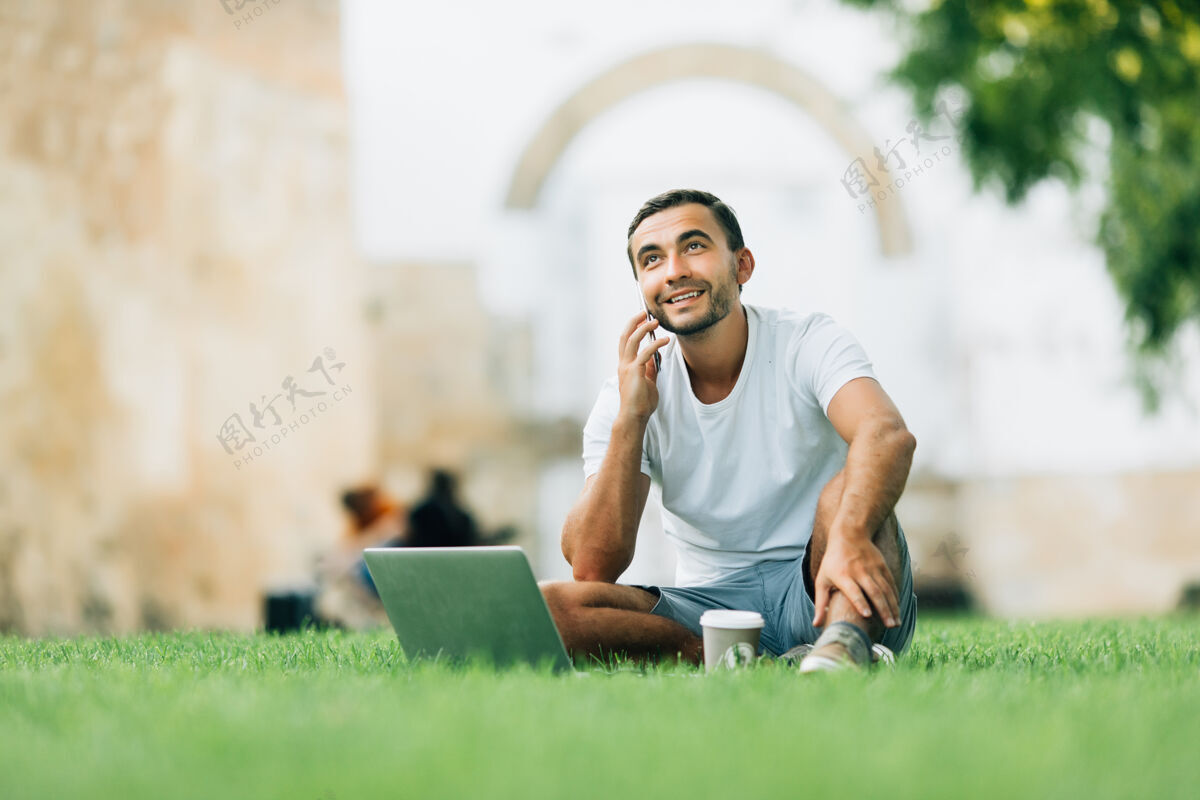 帅气帅哥拿着笔记本电脑坐在城市的草地上讲电话年轻人电话