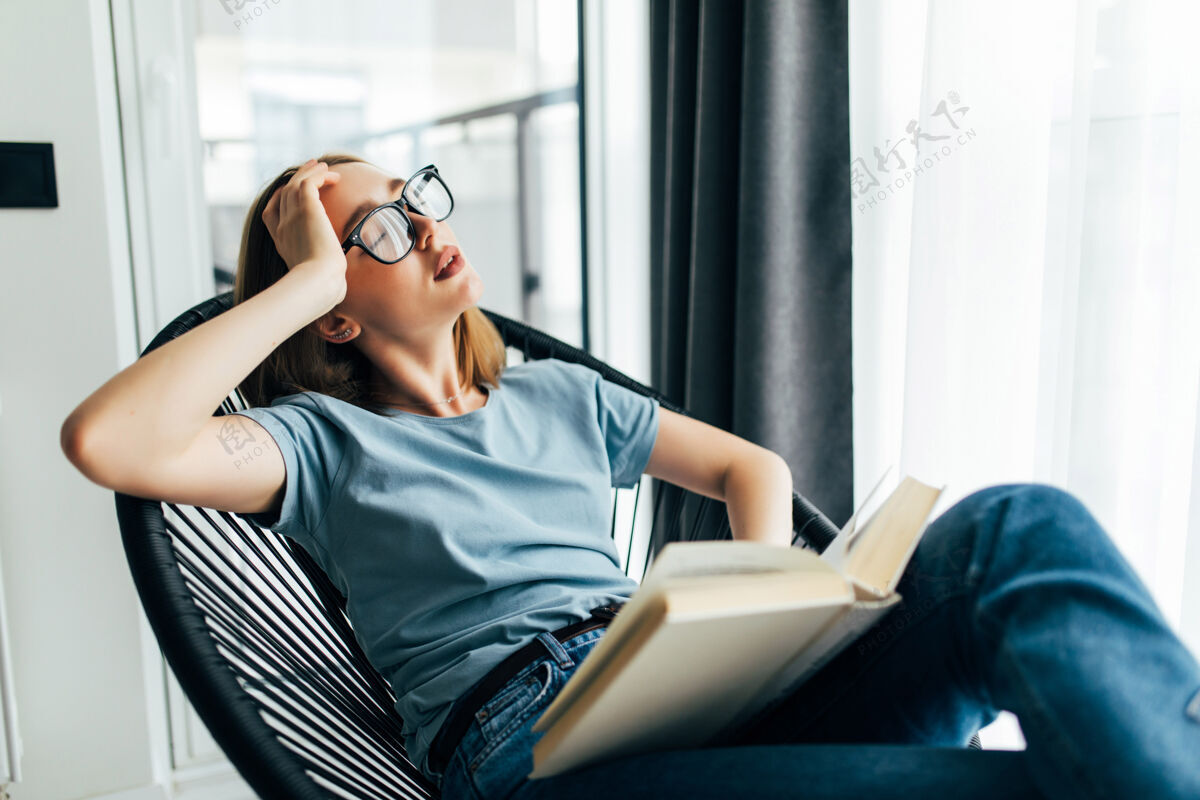 爱好疲倦的年轻女子拿着书睡在家里的躺椅上书房困房间