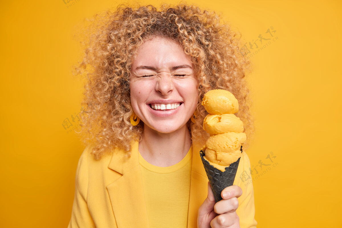 愉悦快乐积极的卷发女人从幸福中闭上眼睛微笑宽大地拿着美味开胃的柠檬味冰淇淋在华夫饼里表达真诚真实的情感单色拍摄满意请圆锥体