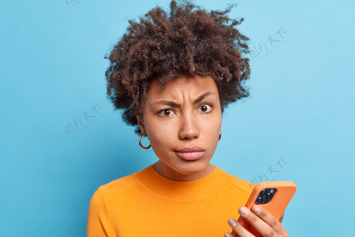 信息不满的非裔美国妇女微笑着脸看着不满的表情手持现代手机在线阅读负面新闻穿着橙色套头衫隔着蓝色的墙科技自负恼怒休闲傻笑