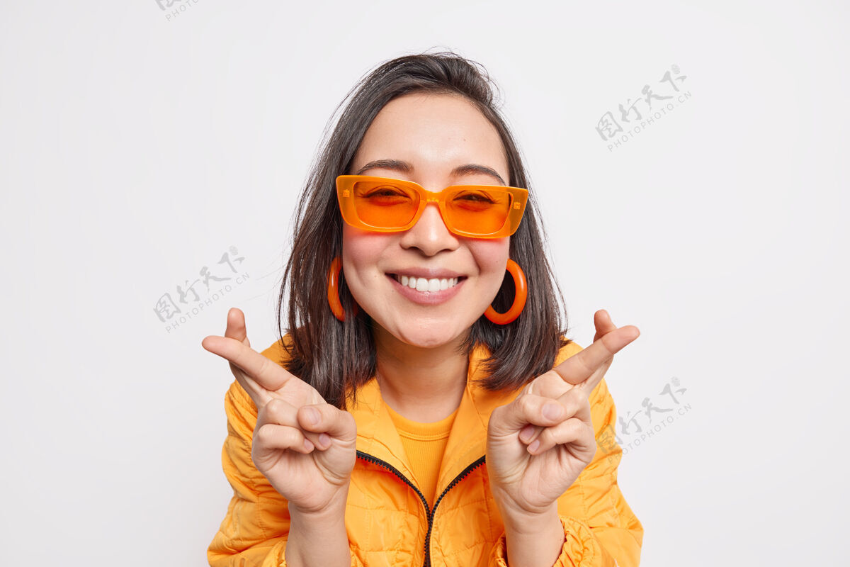 乐观快乐时尚的深色头发的亚洲女人许下愿望祈求梦想成真微笑快乐地戴着时髦的橙色太阳镜耳环和夹克隔着白墙欢呼休闲成人
