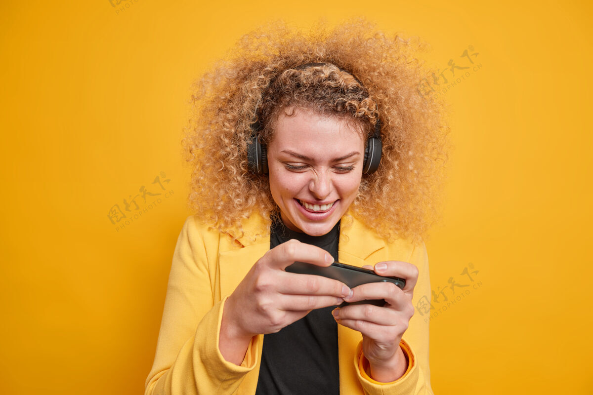 年轻快乐的卷发女人沉迷于电子游戏玩手机使用无线耳机集中在显示器享受游戏装扮正式隔离在生动的黄色墙壁在线互联网微笑