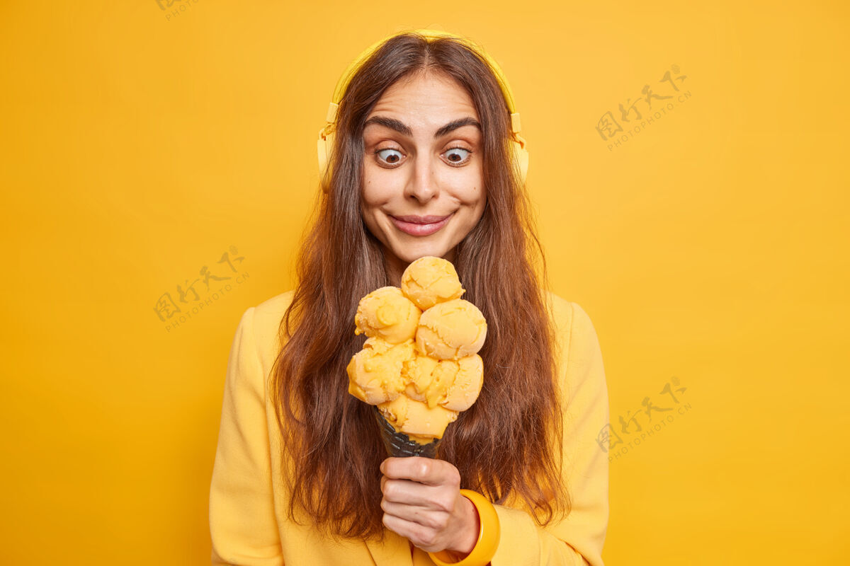 人类图为一位惊讶而快乐的欧洲女性 深色头发 凝视着美味的冰淇淋 饥肠辘辘地吃着 通过立体声无线耳机听音乐 在室内靠着黄色的墙壁摆出姿势高兴美味微笑
