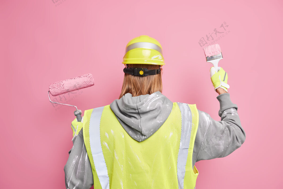 油漆工装修服务理念红发人的后视图使用穿着工作服的建筑设备对着粉色墙壁摆姿势专业的房屋油漆工重新装修房子年轻装修人