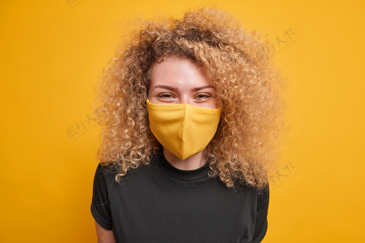 不快乐高兴的卷发黑发女子戴防护面罩 以防止冠状病毒传播穿着休闲黑色t恤表达积极情绪隔离在黄色墙壁检疫时间衣服疾病安全
