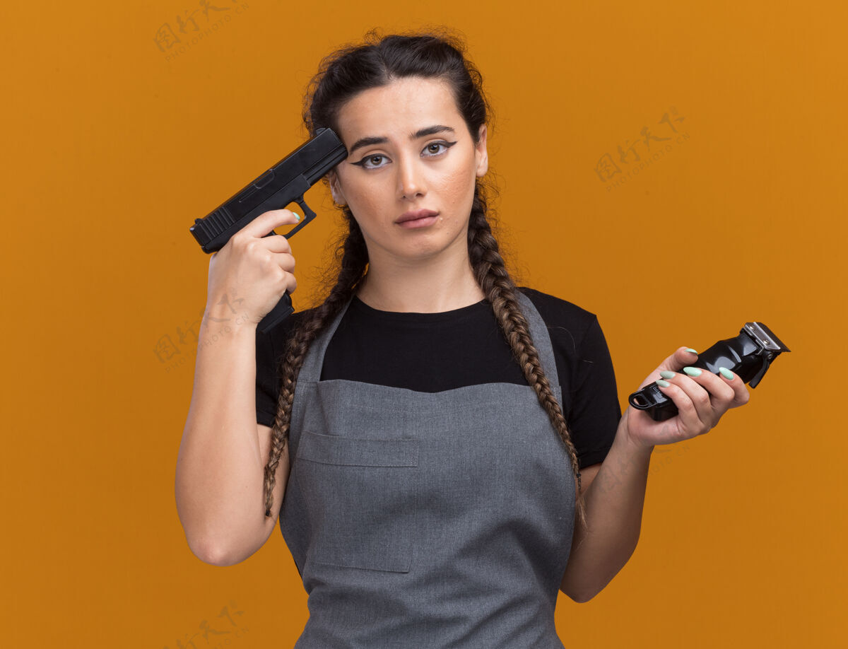 手枪悲伤的年轻女理发师穿着制服拿着理发器 把手枪放在隔离在橙色墙上的太阳穴上姿势女性人