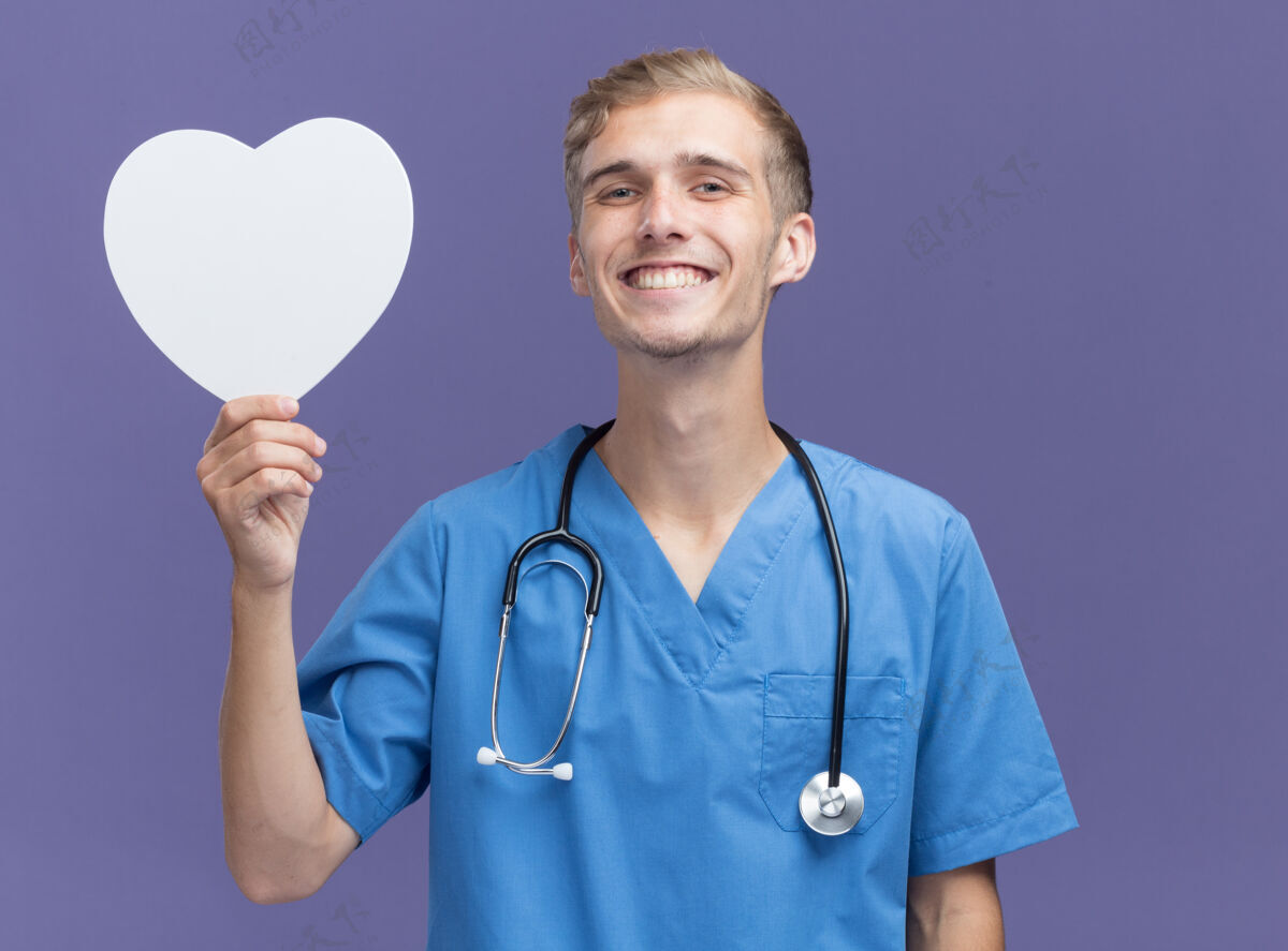 制服微笑的年轻男医生穿着医生制服 手持听诊器 心形盒子孤立在蓝色的墙上感觉脸医生
