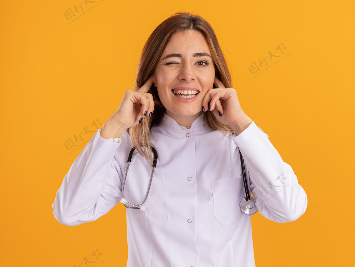 耳朵眨眼的年轻女医生穿着医用长袍 听诊器闭着耳朵隔离在黄色的墙上女性站立眨眼
