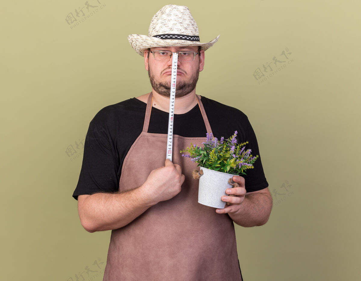 衣服悲伤的年轻男园丁戴着园艺帽 用卷尺在橄榄绿的墙上测量花盆里的花感情人帽子