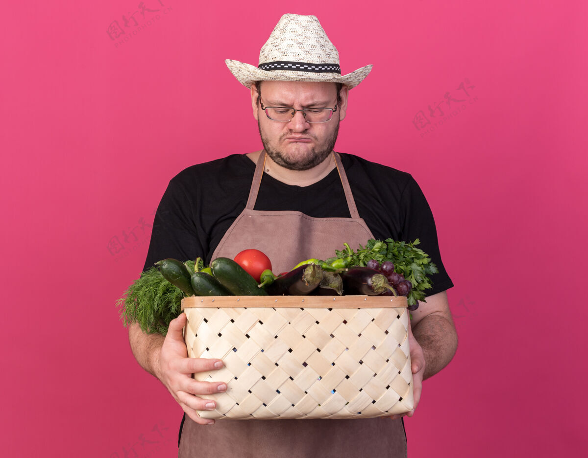 站年轻的男园丁戴着园艺帽 抱着一只孤立在粉红色墙上的菜篮花园表情人