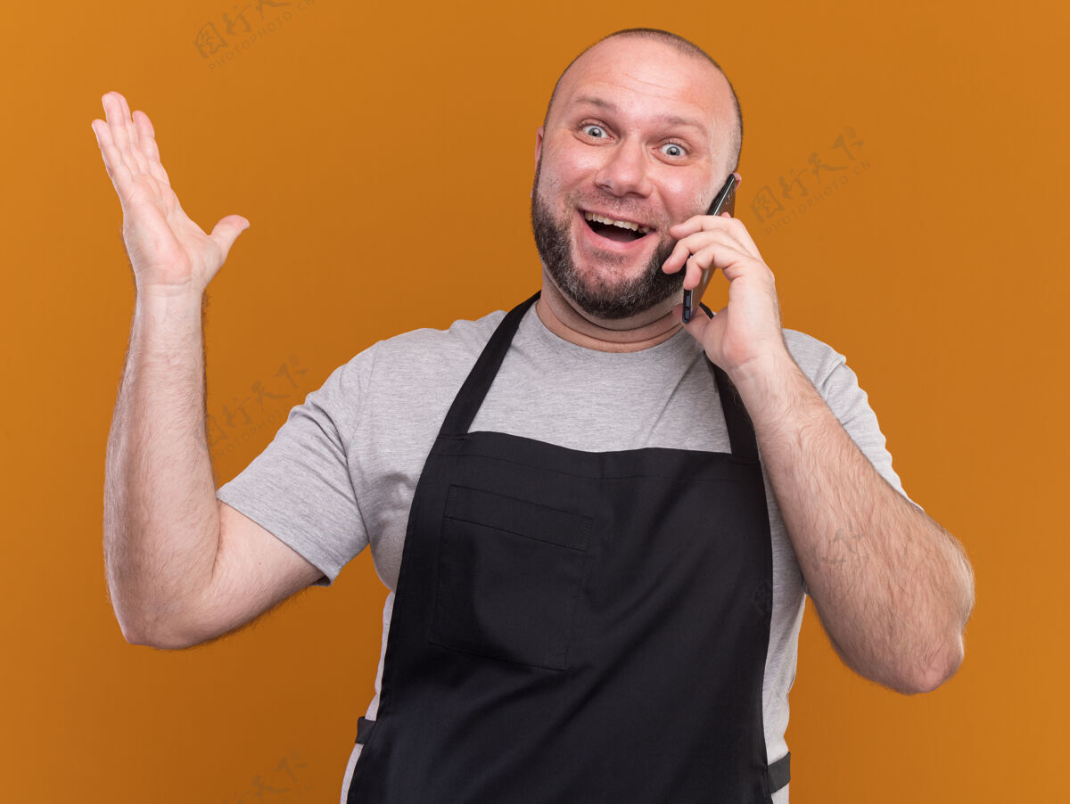 人快乐的中年男理发师穿着制服在电话里说话举起手孤立在橙色的墙上市民感觉快乐