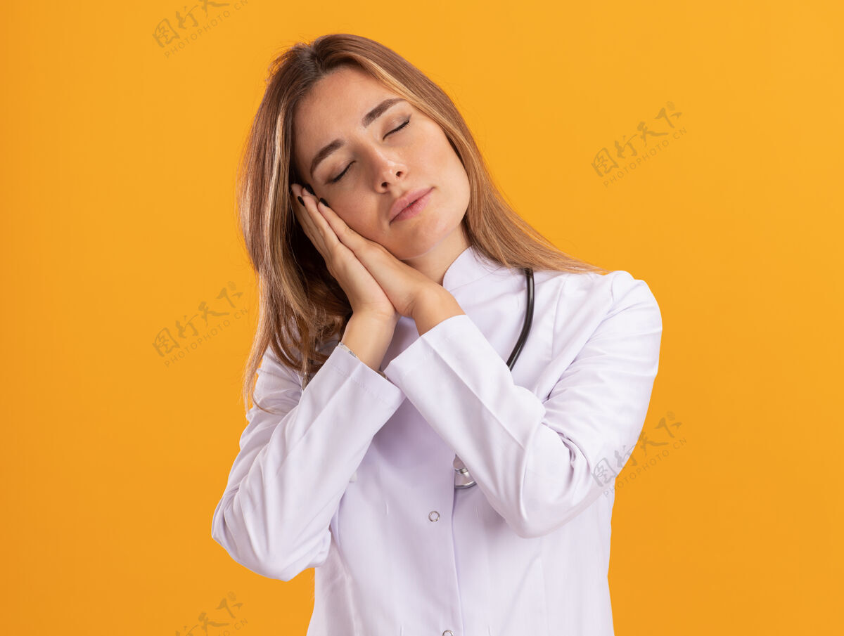 衣服年轻的女医生闭着眼睛 穿着医用长袍 听诊器显示睡眠姿势 隔离在黄色的墙上表情感觉封闭