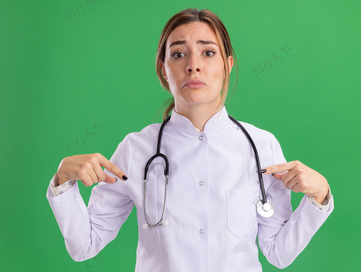 下悲伤的年轻女医生穿着医用长袍 用听诊器指着绿色的墙上隔离下来悲伤站脸