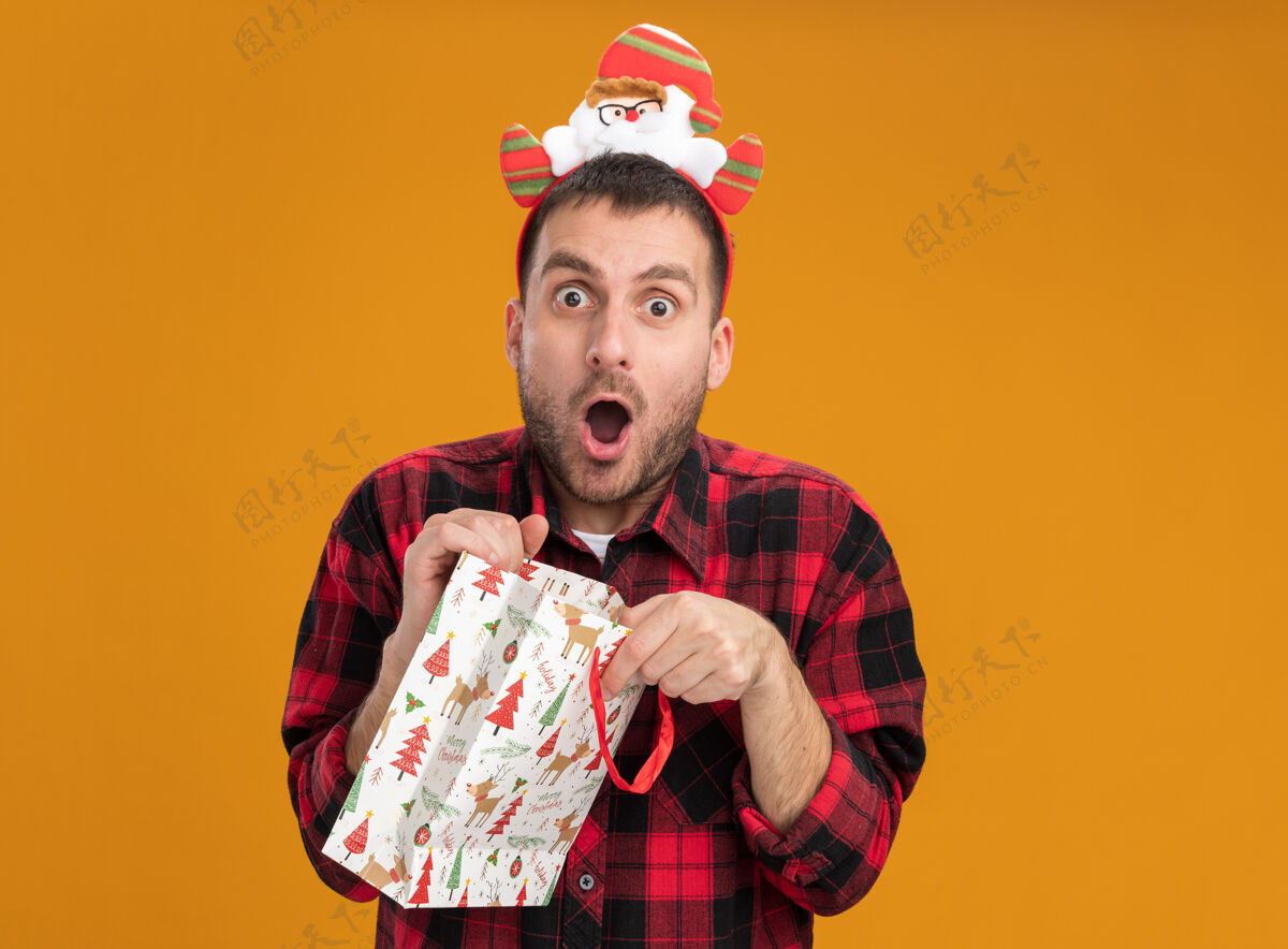白种人惊讶的年轻白种人戴着圣诞老人的头带 拿着圣诞礼物袋 看起来孤立在橙色的墙上复制空间惊喜抱包