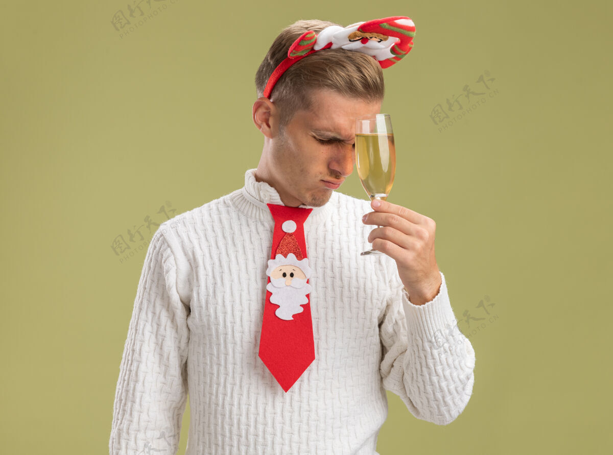 橄榄紧张的年轻帅哥戴着圣诞老人的头带打着领带拿着香槟酒杯抚摸着额头闭着眼睛隔离在橄榄绿的墙上留着复印空间领带圣诞老人年轻