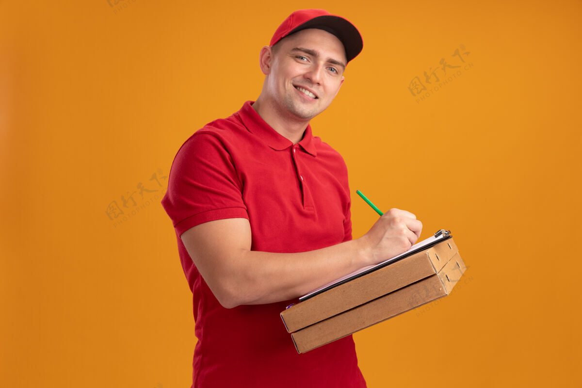 感觉微笑着的年轻送货员穿着制服 戴着帽子 拿着披萨盒 在橘黄色墙上的剪贴板上写着什么人表情人
