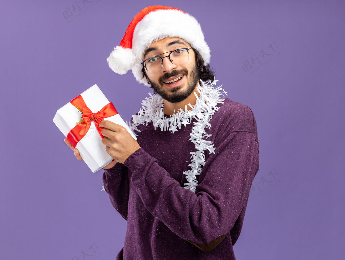 花环微笑的年轻帅哥戴着圣诞帽 脖子上戴着花环 手里拿着礼品盒 孤零零地贴在蓝色的墙上年轻穿脖子
