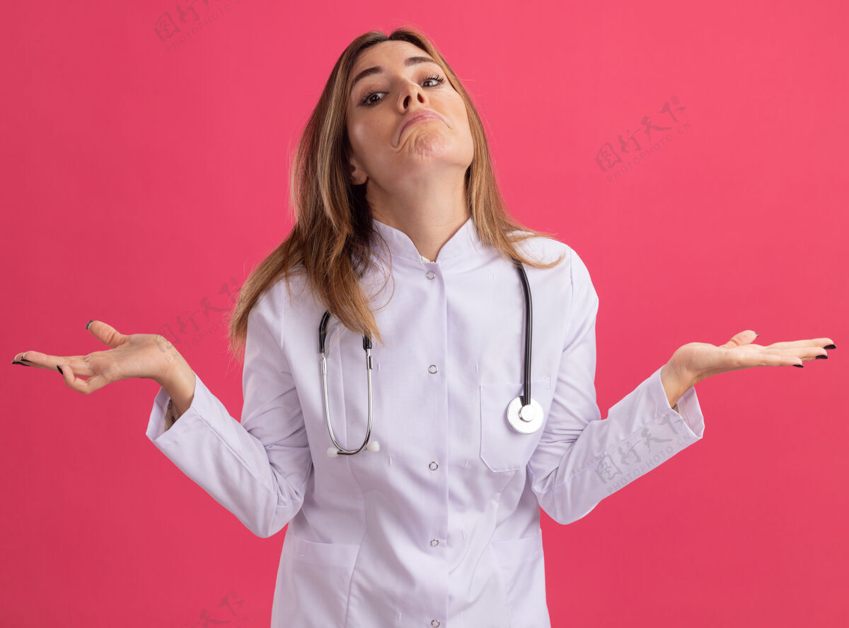 人困惑的年轻女医生穿着医用长袍 听诊器摊开双手隔离在粉红色的墙上人听诊器表情