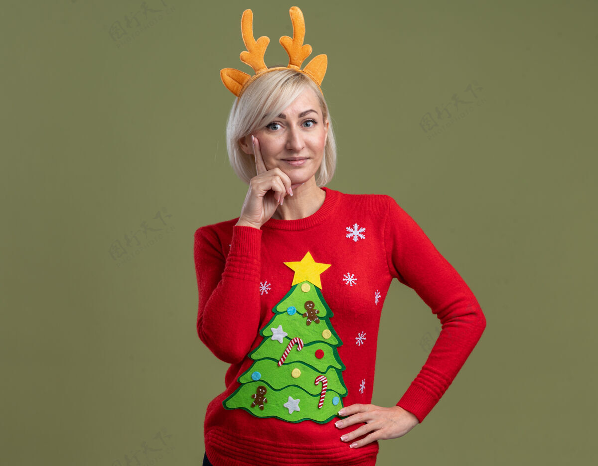 橄榄高兴的中年金发女人穿着圣诞驯鹿鹿角头带和圣诞毛衣看起来保持手放在腰上和下巴上橄榄绿色的墙壁与复制空间隔离腰女人中年