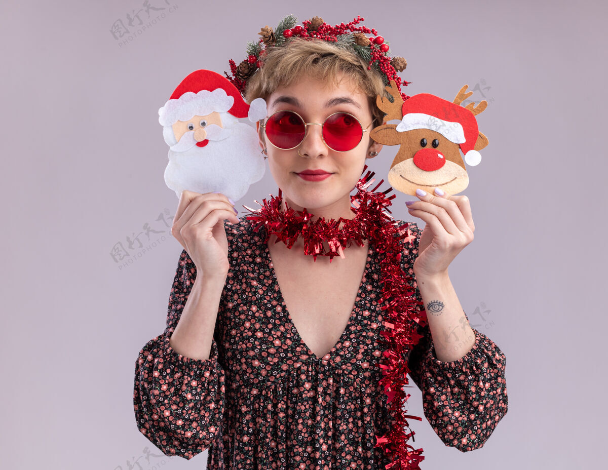 花环高兴的年轻漂亮女孩头戴圣诞花环 脖子上戴着金箔花环 戴着眼镜 头旁拿着圣诞驯鹿和圣诞老人的纸饰品 看着隔离在白色墙上的一面头请女孩