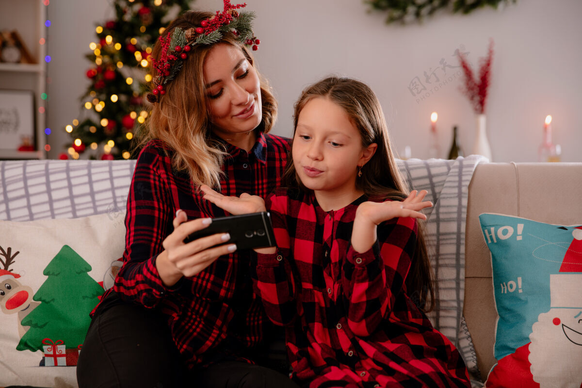 电话高兴的母亲拿着电话 看着困惑的女儿坐在沙发上 在家里享受圣诞节的时光请圣诞节坐着