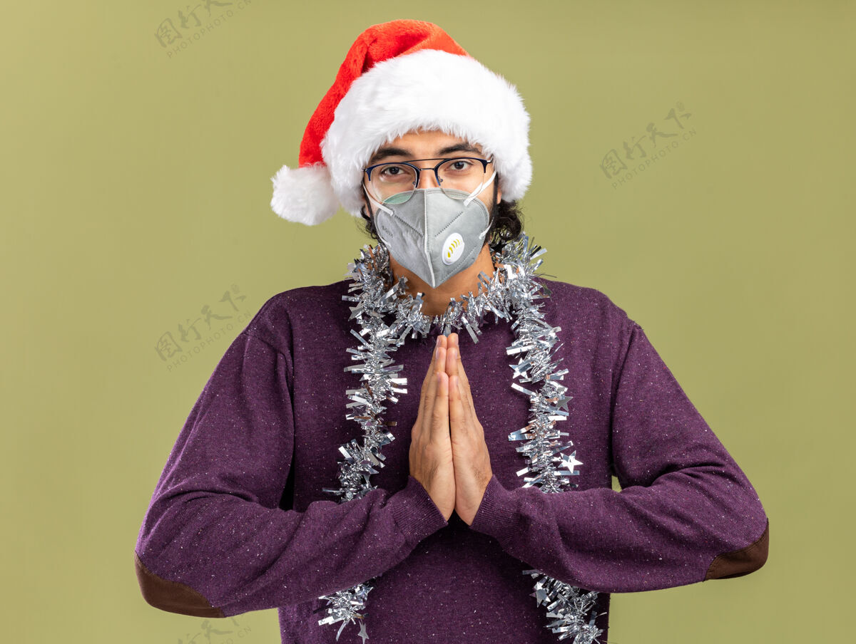 秀高兴的年轻帅哥戴着圣诞帽 戴着医用面具 脖子上戴着花环 在橄榄绿的墙上显示着与世隔绝的祈祷手势橄榄祈祷帅哥