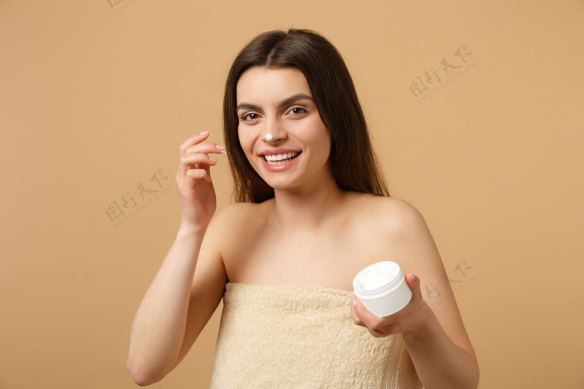 年轻近景半裸体女人完美皮肤裸妆涂抹面霜隔离在米色粉彩墙上手乳液快乐