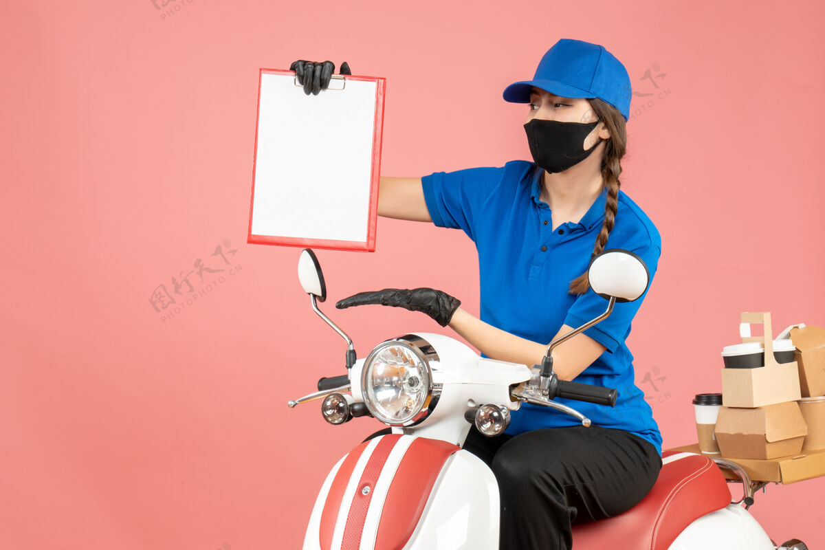 床单顶视图勤劳的快递员女孩戴着医用口罩和手套坐在踏板车上拿着空纸 在柔和的桃色背景下传递订单手套努力工作滑板车