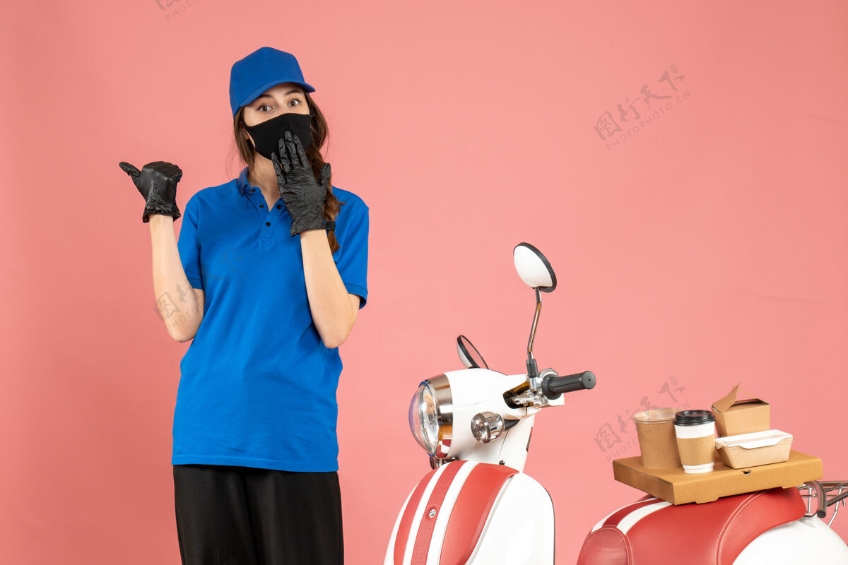 困惑顶视图混乱的快递女孩在医疗面具站在摩托车旁边与咖啡蛋糕上的粉彩桃色背景桃专业站