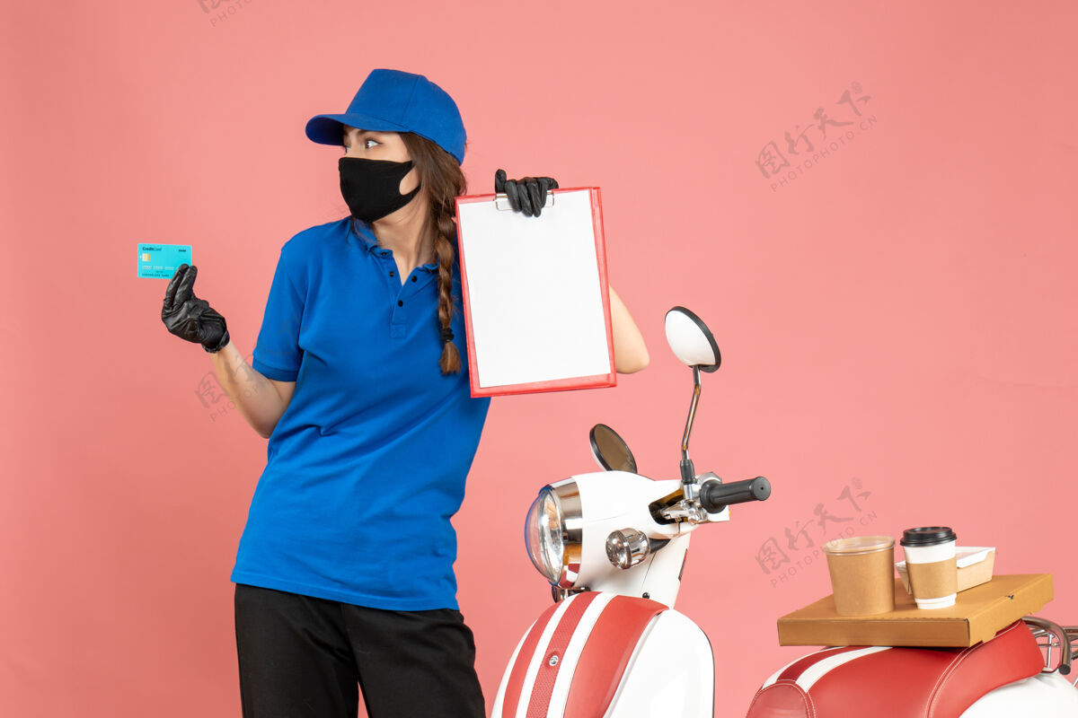 集中俯视图：戴着医用口罩手套的女快递员站在摩托车旁边 摩托车上放着咖啡蛋糕 手里拿着粉色背景的文件和银行卡粉彩摩托车手套