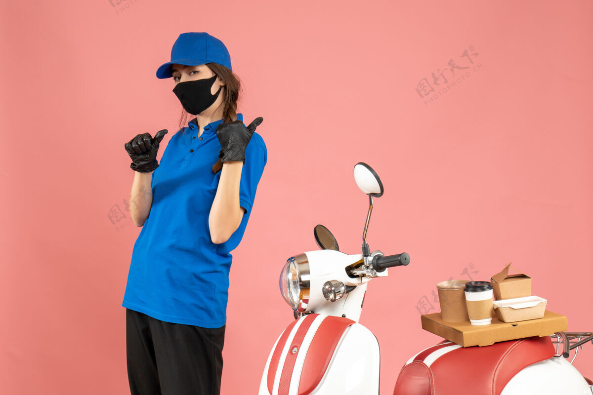 背俯视图：戴着医用面罩手套的信使女孩站在摩托车旁边 上面有咖啡蛋糕 背面是柔和的桃色背景指向男站