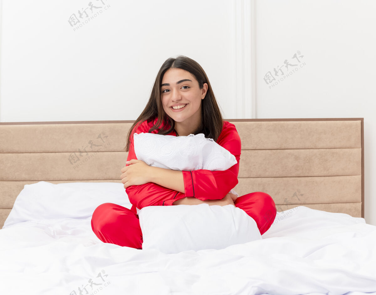 室内穿着红色睡衣的年轻漂亮女人坐在床上抱着枕头看着镜头快乐而积极的微笑在卧室里享受周末看享受床